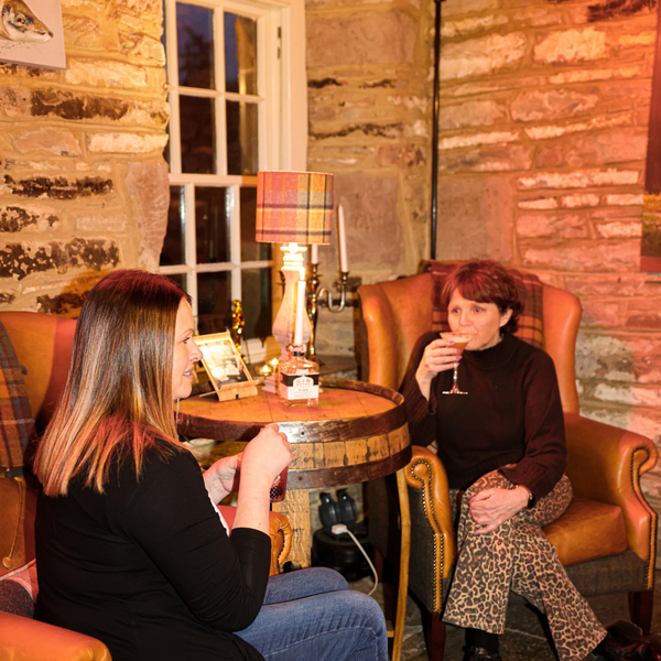 Two women sitting in a cozy room with stone walls, one holding a glass.