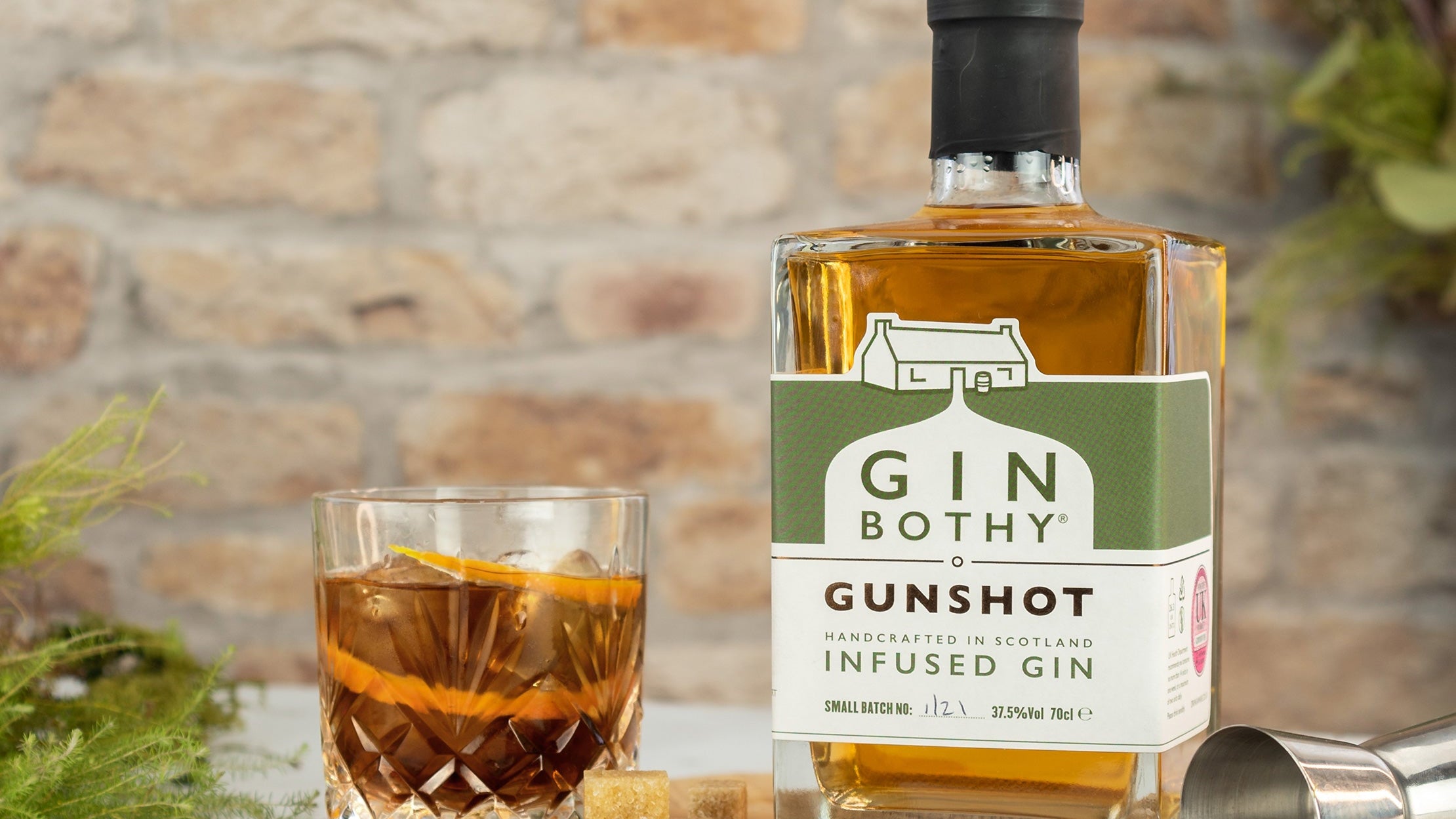 Bottle of Gin Bothy Gunshot Gin with a glass of cocktail on a wooden board against a stone wall background.