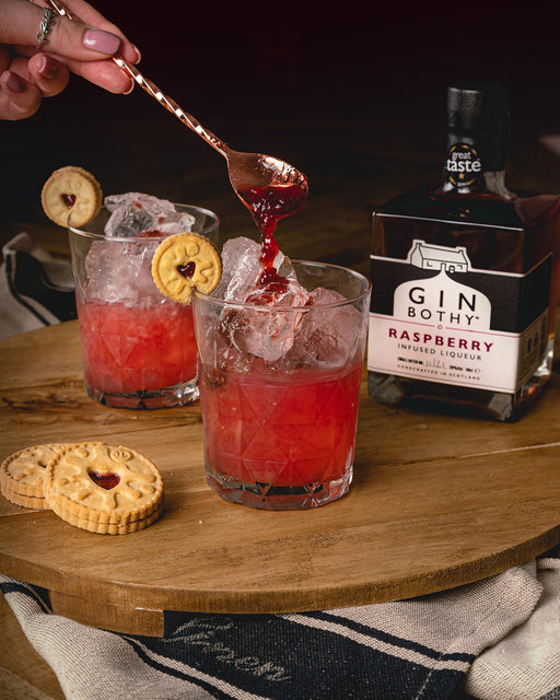 Gin cocktail being garnished with a raspberry syrup, with a bottle of Gin & Tonic Raspberry in the background.